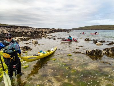A sea kayaker pulls her boat out of the water on to a rocky shore, other kayakers approach in the background